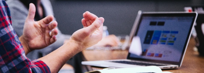 photo of a man making a hand gesture while talking, sitting in front of a laptop. 