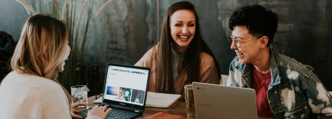 photo of four people looking at a laptop screen in a library or coffee shop. 