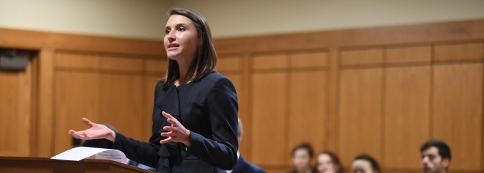 Picture of a law student speaking at a podium in a courtroom. 