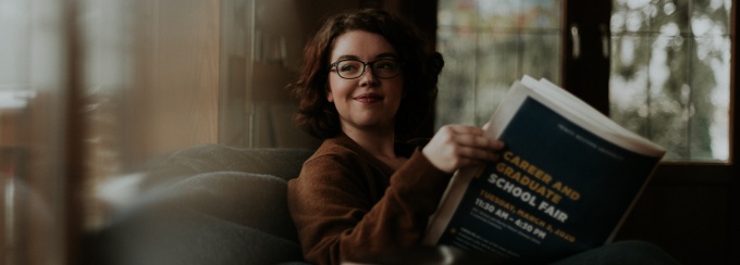 Women sitting on the couch, looking out the window, the sun is shining through. She is holding a newspaper that reads "Careers and Graduate School Fair". 