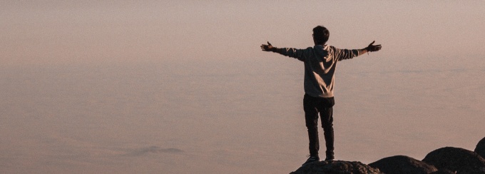 photo of the back of a person raising hands while standing on a mountain with other mountains in view. 