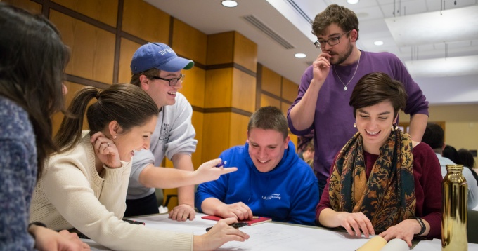 Six University at Buffalo students gathering around a tabling smiling and talking, while looking at paperwork. 