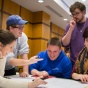 Six University at Buffalo students gathering around a tabling smiling and talking, while looking at paperwork. 