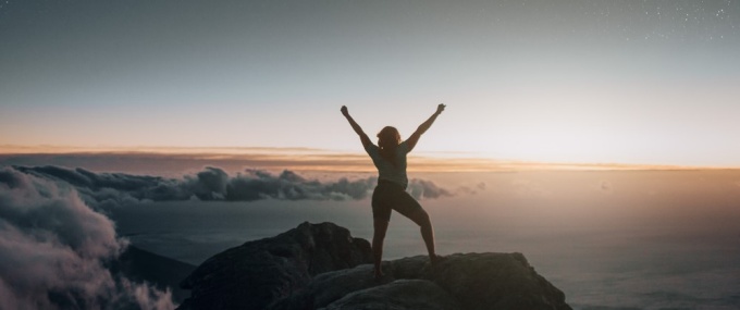 women standing proudly atop a mountain, above the clouds, during sunrise. 