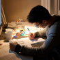 Person seated at a desk studying under warm lighting, writing notes in a notebook while referencing an open book and a computer monitor. The desk is cluttered with study materials, including pens, notebooks, and a mug, suggesting focused preparation or exam studying. 
