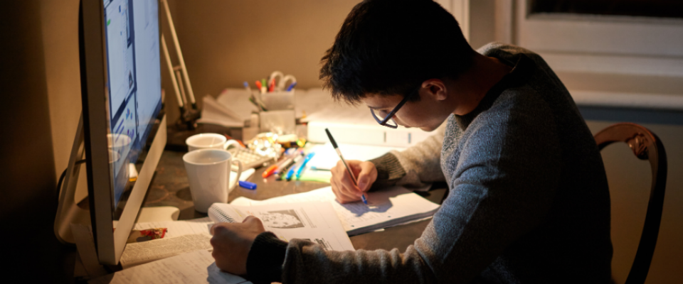 Person seated at a desk studying under warm lighting, writing notes in a notebook while referencing an open book and a computer monitor. The desk is cluttered with study materials, including pens, notebooks, and a mug, suggesting focused preparation or exam studying. 