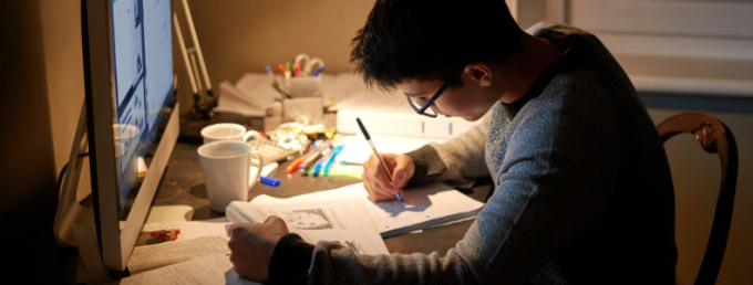 Person seated at a desk studying under warm lighting, writing notes in a notebook while referencing an open book and a computer monitor. The desk is cluttered with study materials, including pens, notebooks, and a mug, suggesting focused preparation or exam studying. 