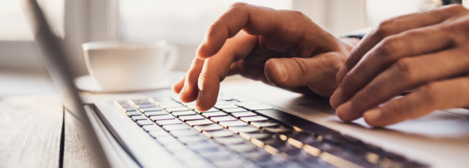 Close-up of hands typing on a laptop keyboard with a blurred white coffee cup in the background. 