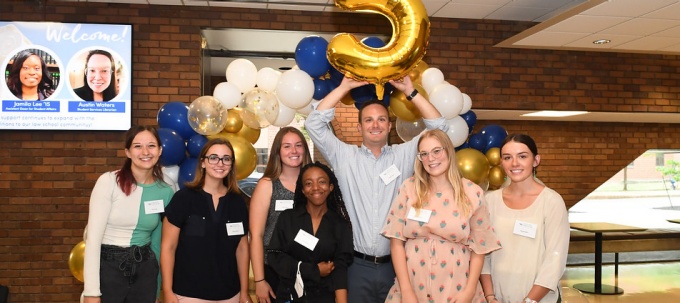 a group of students standing in O'Brian Hall's lobby, one student holding up a balloon in the shape of the number 5. 