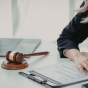 Wooden gavel and scales of justice on a desk beside a laptop, with a person&rsquo;s hands reviewing documents. 