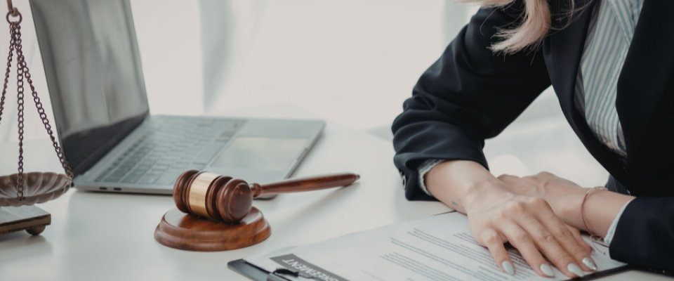 Wooden gavel and scales of justice on a desk beside a laptop, with a person&rsquo;s hands reviewing documents. 