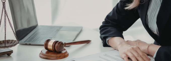Wooden gavel and scales of justice on a desk beside a laptop, with a person’s hands reviewing documents. 