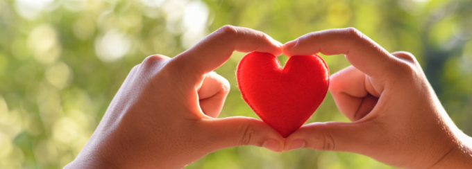 Two hands hold a small red heart between the fingers, forming a heart shape, with a softly blurred green outdoor background. 
