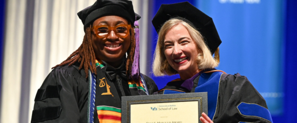Award recipient and faculty member in academic regalia display a framed certificate during a law school commencement ceremony. 