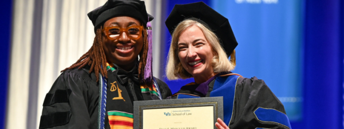 Award recipient and faculty member in academic regalia display a framed certificate during a law school commencement ceremony. 