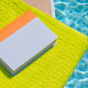 Closed book resting on a bright green towel beside a swimming pool, with sunlight reflecting on the water's surface. 