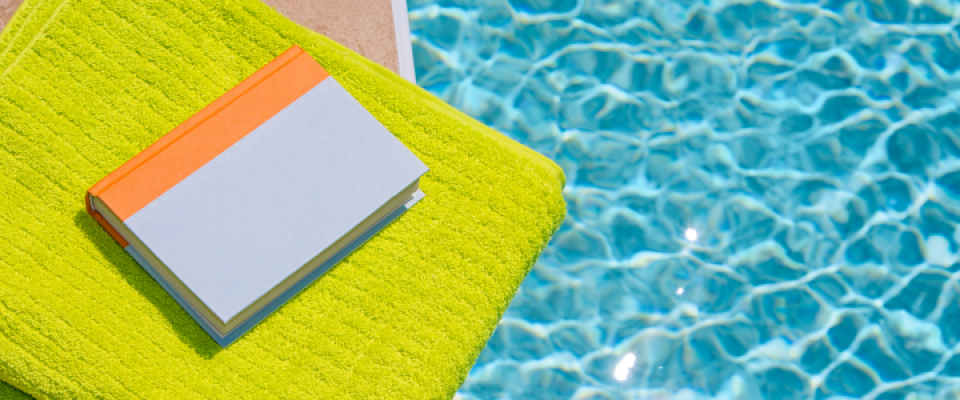 Closed book resting on a bright green towel beside a swimming pool, with sunlight reflecting on the water's surface. 