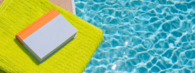 Closed book resting on a bright green towel beside a swimming pool, with sunlight reflecting on the water's surface. 