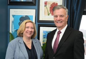 Two adults stand side by side in an office, facing the camera. One person on the left wears a light blue textured blazer over a dark top with a pearl necklace, and the person on the right wears a dark pinstripe suit with a white shirt and red patterned tie. Behind them is a blue wall with several framed artworks, including a poster that reads “Brown v. Board of Education 30 Years Later: ‘The Politics of Excellence,’” along with floral and landscape prints. A large window with dark curtains appears on the right, and potted plants are visible near the floor. 