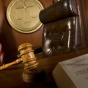 A low-angle view of a courtroom scene featuring a wooden judge&rsquo;s bench with a leather chair behind it, a wooden gavel resting on the desk in the foreground, and an open book partially visible. An American flag stands to the side, and a circular wall emblem with scales of justice is mounted behind the chair. 