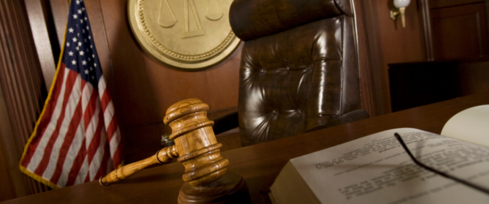 A low-angle view of a courtroom scene featuring a wooden judge&rsquo;s bench with a leather chair behind it, a wooden gavel resting on the desk in the foreground, and an open book partially visible. An American flag stands to the side, and a circular wall emblem with scales of justice is mounted behind the chair. 