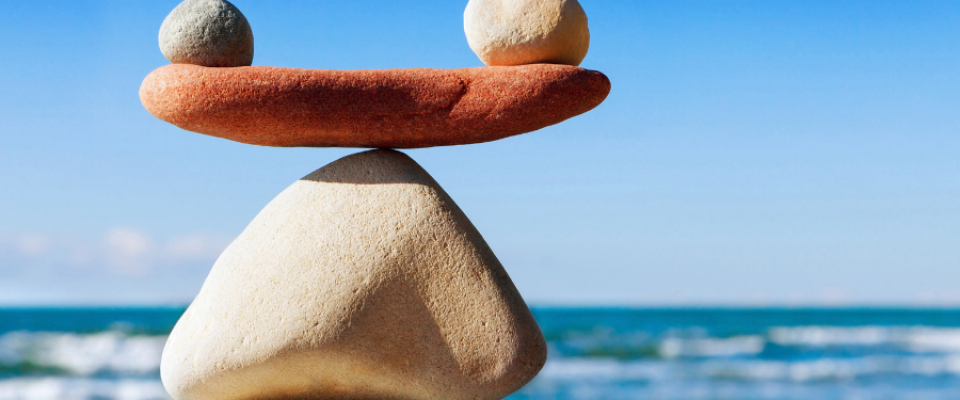 Three stones balanced in a minimalist arrangement against a clear blue sky and ocean backdrop. A large triangular stone forms the base, topped by a flat reddish stone with two round stones of different colors resting on each end, symbolizing balance and harmony. 