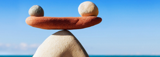 Three stones balanced in a minimalist arrangement against a clear blue sky and ocean backdrop. A large triangular stone forms the base, topped by a flat reddish stone with two round stones of different colors resting on each end, symbolizing balance and harmony. 