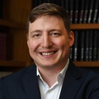 man smiling, standing in a room with books on bookshelves. 