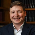 man smiling, standing in a room with books on bookshelves. 