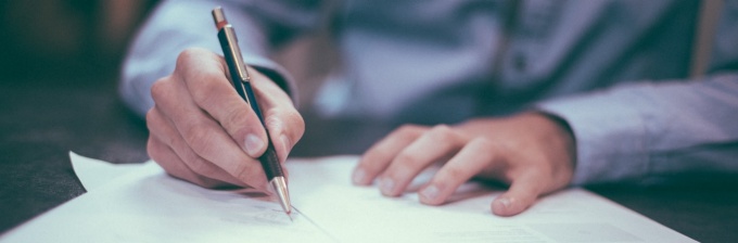 photo of man with pen in his hand, writing on a piece of paper. 