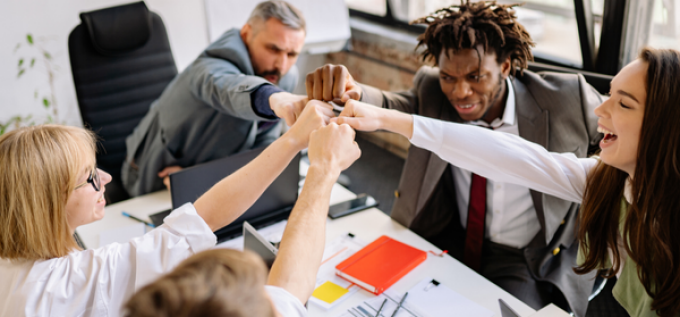 Several people seated around a table in a modern office space extend their hands into the center, touching fists in a group gesture. Laptops, notebooks, and office chairs are visible around the table, suggesting a collaborative work meeting. 