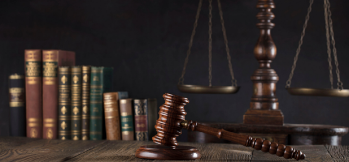 Wooden judge’s gavel resting on a desk with a stack of law books and a balance scale in the background. 