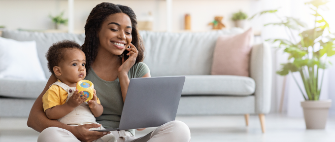 woman sitting on the floor with a baby on her lap, holding a laptop and smiling as she talks on her mobile phone. 