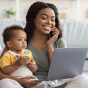 woman sitting on the floor with a baby on her lap, holding a laptop and smiling as she talks on her mobile phone. 