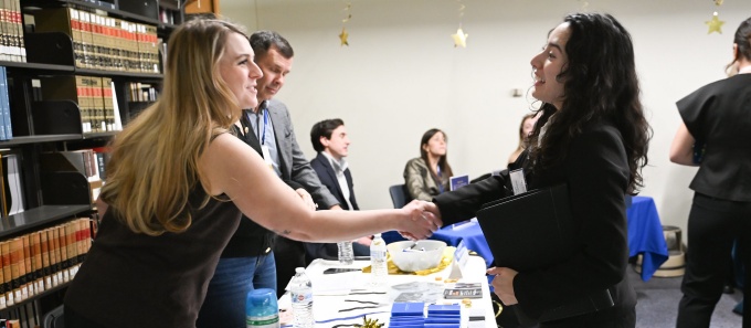 Two attendees shake hands across a career fair table inside a library, with informational materials, branded items, and bookshelves visible behind them while other participants sit and stand nearby. 