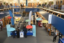 Wide interior view of a library atrium during a career fair, showing multiple information tables, standing banners, colorful open umbrellas, and many attendees gathered across two floors with bookshelves in the background. 