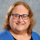 woman wearing blue sweater, glasses, smiling, blue and grey background. 