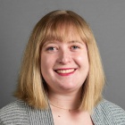 woman wearing sweater, flowery top, standing in front of grey background, smiling. 