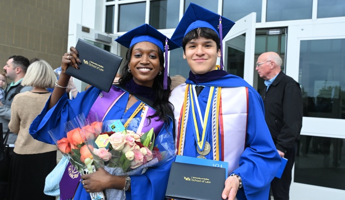 Two graduates in blue caps and gowns stand outside a building holding their diploma covers. One holds a bouquet of roses, and both wear honor cords and stoles as people gather in the background. 