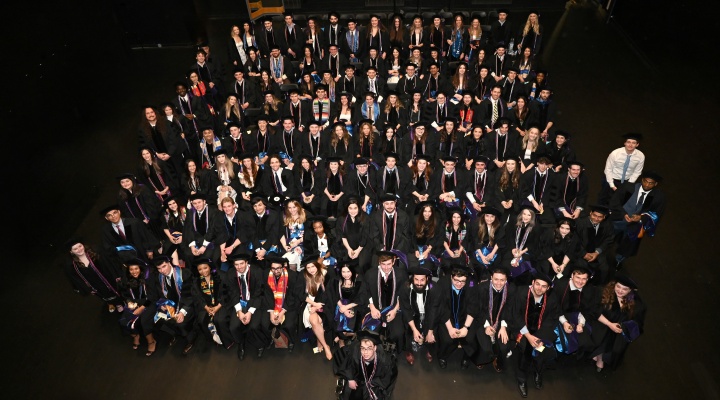 Large group of graduating students in black caps and gowns seated in tiered rows inside a dark auditorium, viewed from above. Many wear honor cords or stoles in various colors, and several hold programs or small items while facing the camera for a formal group photo. 