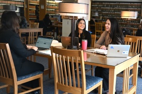 Three female students sitting at a table in a library with laptop computers. 
