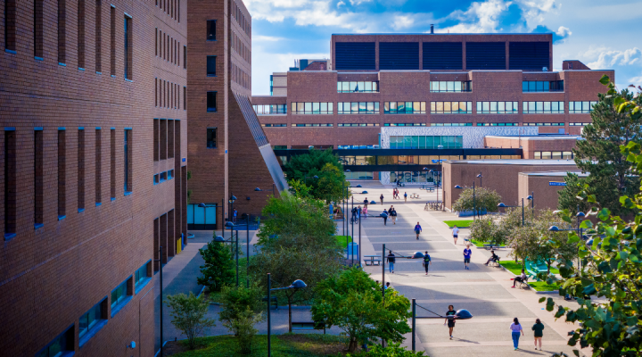 View of a university campus walkway between academic buildings, with students walking and gathering along a tree-lined pedestrian corridor under a partly cloudy sky. 