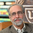 Binder seated in a chair inside a law library, with bookshelves and a large carved university seal visible in the background. 