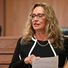 A person wearing a black jacket with white trim and a pearl necklace is standing in a courtroom, holding a printed document with highlighted text. The background shows a wooden judge’s bench and two black leather chairs. 
