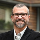 Portrait of a person wearing glasses, a dark blazer, and a white collared shirt. 