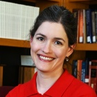 Close‑cropped professional portrait of a person wearing a red collared top, photographed indoors in front of wooden bookshelves filled with books, suggesting an office or library setting. 