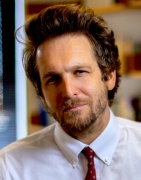 Close up portrait of Linden-Retek wearing a white shirt and red patterned tie, standing indoors with shelves and books softly blurred in the background. 