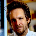 Close up portrait of Linden-Retek wearing a white shirt and red patterned tie, standing indoors with shelves and books softly blurred in the background. 