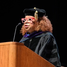 woman wearing graduation regalia, speaking into a podium. 