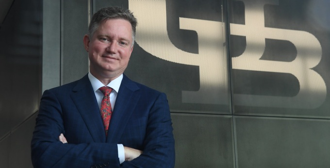man standing, arms crossed, in front of a sign that says UB. 